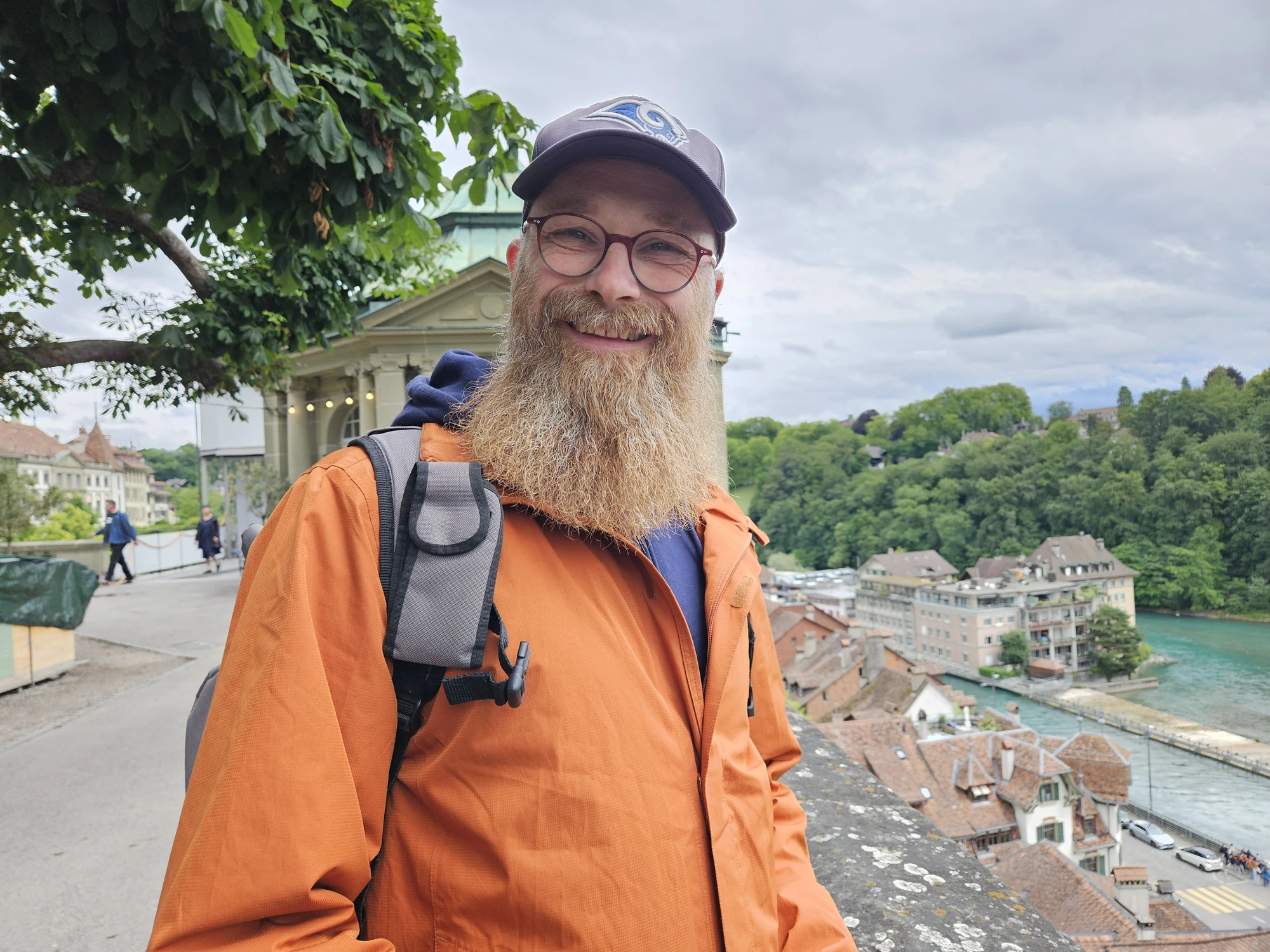 Thorsten Weber im Freien, lächelnd, mit Rucksack vor einer Stadt- und Flusslandschaft.